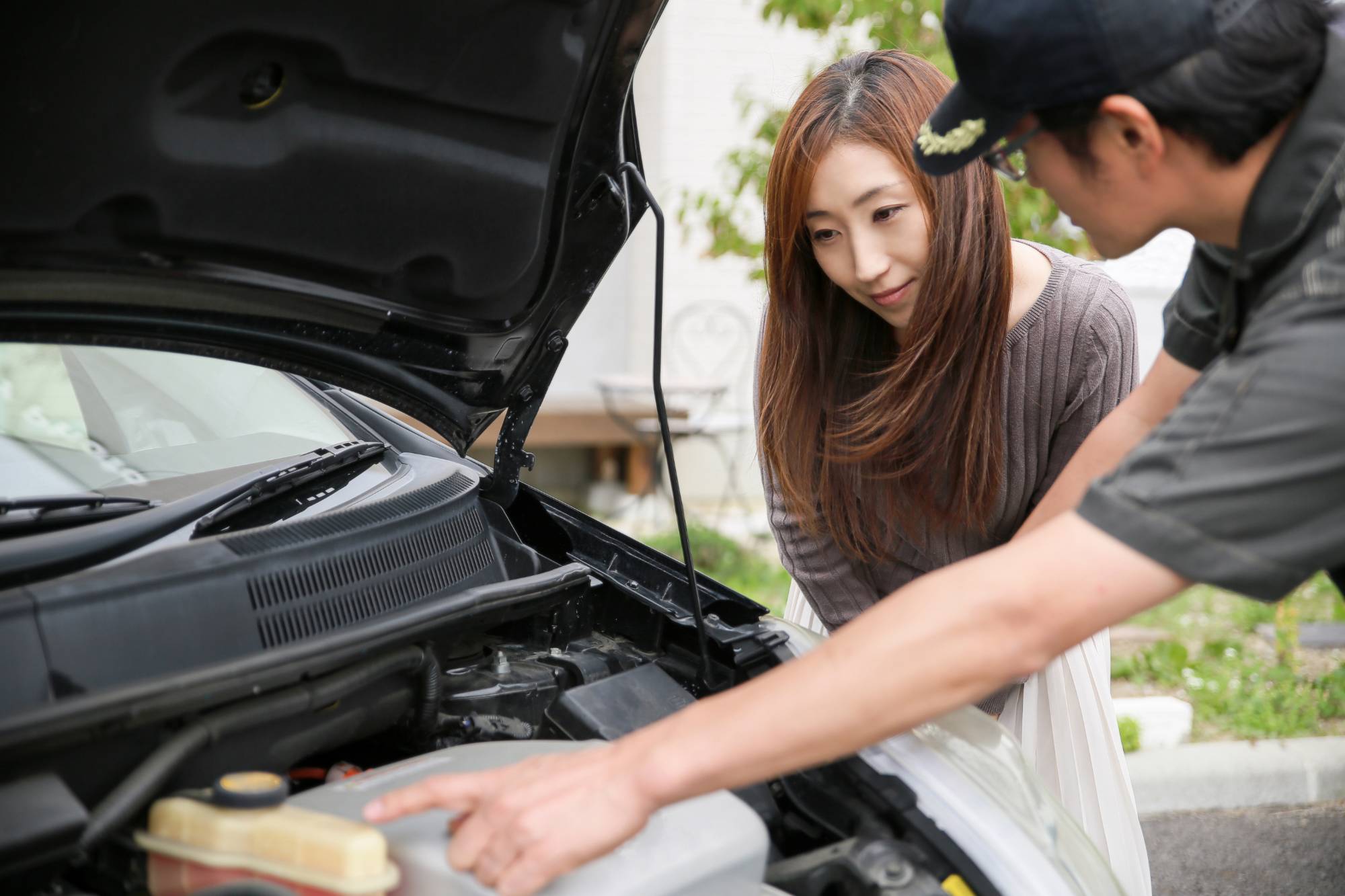 黒い車のボンネットを開けてエンジンの説明をする整備士と見守る女性